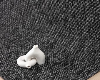 May include: A dark gray woven rug with a textured, heathered appearance featuring a dense, uniform pattern. Resting on the surface is a small, white, abstract ceramic sculpture composed of two interconnected, rounded, hollow forms.