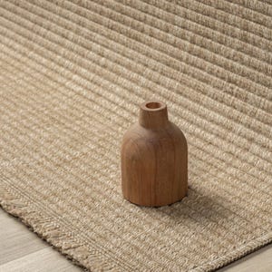 May include: A small, wooden vase with a rounded body and a narrow neck sits on a beige, textured rug. The rug has a ribbed pattern and a fringed edge. The vase is made of light brown wood.