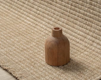 Puede incluir: Un pequeño jarrón de madera de color marrón claro, liso, con cuello estrecho y cuerpo redondeado, colocado sobre una alfombra beige texturizada de canalé que presenta un borde tejido y un borde ligeramente deshilachado.
