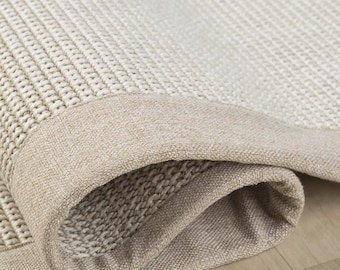 Puede incluir: Una alfombra tejida con un centro texturizado de color blanco roto con un patrón de rejilla apretado. La alfombra está rematada con un borde de tela beige liso y sólido que envuelve el borde, mostrando una esquina doblada.