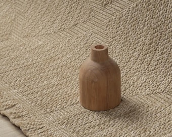 May include: A small, light brown wooden vase with a narrow neck and rounded body sits on a textured, beige woven rug. The rug features a prominent herringbone pattern and a thick, knotted border along the edge, creating a tactile surface.