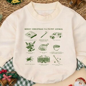 May include: Cream-colored baby bodysuit with long sleeves. The front features green illustrations and the text "MERRY CHRISTMAS YA FILTHY ANIMAL." The design includes images of a pizza, a toothbrush, a van, and more.