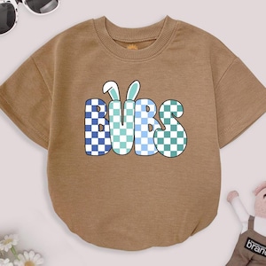 May include: A tan short-sleeved baby bodysuit with the word "BUBS" in a checkered pattern of blue, light blue, and green. The word "BUBS" has bunny ears above it. Sunglasses and a stuffed animal are in the background.
