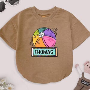 May include: A tan short-sleeved shirt featuring a colorful beach ball graphic with the name "THOMAS" in a rectangular box. The beach ball design includes sections of yellow, orange, green, purple, and pink. The shirt is a casual style.