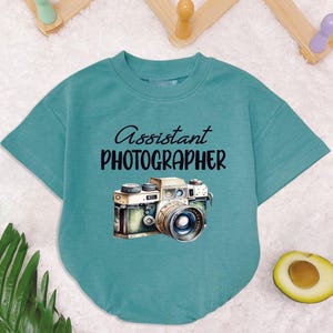 May include: A teal blue t-shirt with short sleeves and a crew neck. The shirt has a graphic of a vintage camera and the text "Assistant Photographer".