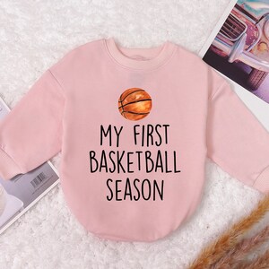 May include: Pink long-sleeved sweatshirt with a basketball graphic and the text "My First Basketball Season".