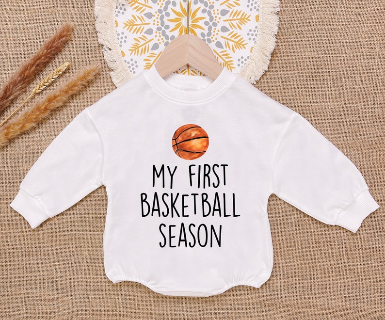 May include: White long-sleeved onesie with a basketball graphic and the text "My First Basketball Season!"