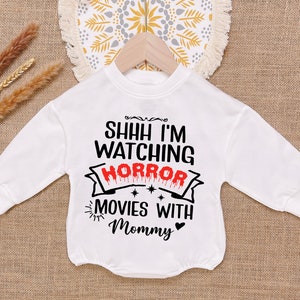 May include: White long-sleeve shirt with black text that reads "Shhh I'm watching horror movies with mommy" with a red dripping heart.