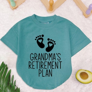 May include: A teal blue short-sleeved t-shirt with a black graphic of two footprints with hearts inside. The text "Grandma's Retirement Plan" is printed below the footprints.