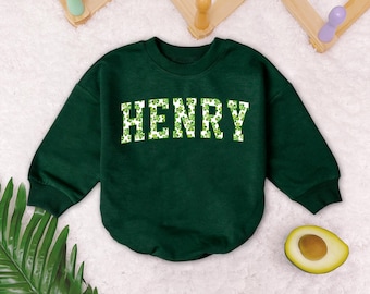 May include: A dark green sweatshirt with the name "HENRY" in white letters filled with green shamrock patterns. The sweatshirt has long sleeves and a round neckline. An avocado half is in the bottom right corner.