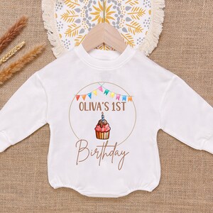 May include: White long-sleeve onesie with a brown circle design. The circle has colorful flags, the text "OLIVA'S 1ST", a cupcake with a lit candle, and the text "Birthday".