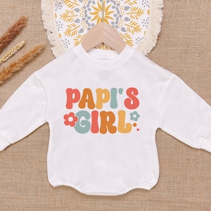 May include: White long-sleeve shirt with the text "Papi's Girl" in colorful retro-style lettering with flowers.