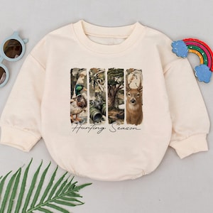 May include: Cream-colored long-sleeved sweatshirt with a graphic featuring a duck, binoculars, a tree, and a deer. The text "Hunting Season" is printed below the images. Sunglasses and a rainbow-shaped toy are on the sides.