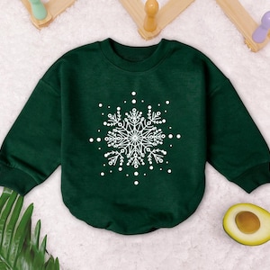 May include: A dark green sweatshirt with a white snowflake design. The snowflake is detailed with intricate lines and swirls.