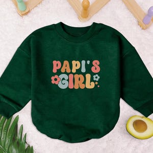May include: A dark green sweatshirt with the text "Papi's Girl" in colorful retro lettering and a flower graphic.