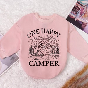 May include: A light pink baby sweatshirt with long sleeves. The front features a black graphic of a mountain scene with a tent, trees, and the text "ONE HAPPY CAMPER".