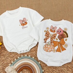 May include: Two white baby rompers. One has a pumpkin and bow design with the name "Olivia". The other has a turkey, bows, and pumpkin design. Both have short sleeves and snap closures.