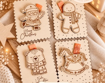 May include: Four rectangular wooden cards with scalloped edges, each featuring a laser-engraved holiday design: a Santa Claus with Merry Christmas text, a gingerbread person, a bear with a scarf and Frohe Weihnachten text, and a rocking horse.
