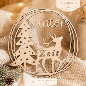 Könnte beinhalten: Ein hölzerner Weihnachtsschmuck mit einem Baum und einem Hirsch in einem kreisförmigen Rahmen. Das Wort "Winter" steht oben und "Zeit" auf dem Hirsch. Der Schmuck ist aus hellem Holz gefertigt.