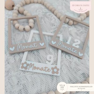 May include: Three rectangular photo frame markers in beige and white, featuring the German text 'Monat' or 'Monate' and numbers 9 and 12. The frames are decorated with small hearts or flowers and are arranged with a string of wooden beads.