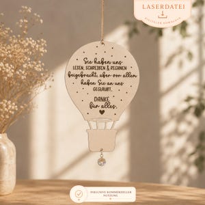 May include: A light-coloured wooden hot air balloon ornament hanging by a string. The surface features laser-engraved German text: Sie haben uns LESEN, SCHREIBEN & RECHNEN beigebracht, aber vor allem haben Sie an uns GEGLAUBT. DANKE für alles. A small crystal bead hangs from the bottom.