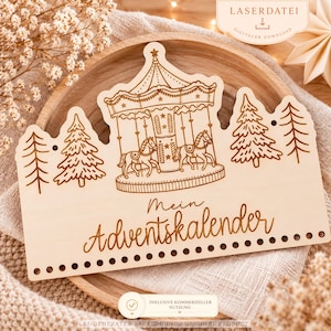Puede incluir: Una placa de madera clara con un grabado láser de un carrusel y árboles de pino. El texto dice Mein Adventskalender. El borde inferior tiene una fila de pequeños agujeros circulares para colgar objetos decorativos.