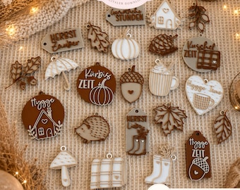 May include: A collection of decorative autumn-themed ornaments in various shapes including pumpkins, leaves, houses, mushrooms, acorns, and boots. The pieces feature brown, white, and beige colours with engraved text and patterns, designed as 3D print files.