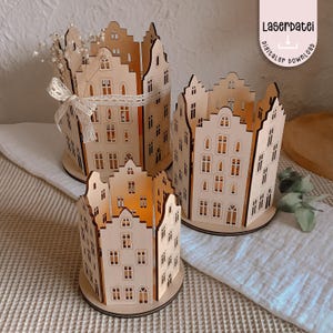 Könnte beinhalten: Drei hölzerne Kerzenhalter in Form von Gebäuden mit mehreren Fenstern und einer hellen Oberfläche. Ein Halter ist mit einem Spitzenband und Trockenblumen verziert. Die Kerzenhalter stehen auf einem Holzsockel.