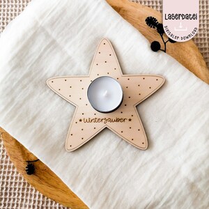Può includere: Un portacandele in legno a forma di stella con una candela tealight bianca al centro. La stella è decorata con piccoli punti marroni e il testo "*Winterzauber*" è inciso sul fondo.