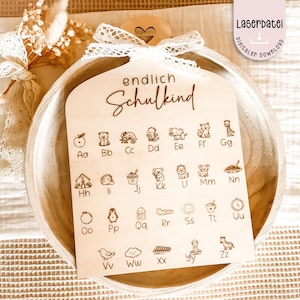 May include: A wooden sign with the words "endlich Schulkind" and an alphabet with animal illustrations. The sign is decorated with a white lace bow and a small wooden heart. The sign also has the text "Laserdatei digitaler download".