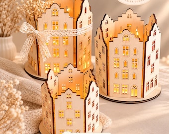 Può includere: Tre strutture in legno chiaro a forma di case in miniatura con finestre e porte ritagliate. Le strutture sono illuminate dall'interno da luci calde. Una casa presenta un nastro decorativo in pizzo bianco legato attorno.
