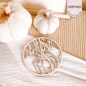 Könnte beinhalten: Helle Holz-Kreisdekoration mit der Aufschrift "Happy Herbst" und einem Kürbis-Design. Die Dekoration liegt auf einer weißen Oberfläche. Zwei weiße Kürbisse befinden sich im Hintergrund.
