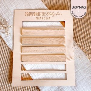 Może przedstawiać: Jasna drewniana tablica ekspozycyjna z napisem "GRAVURHALTER Klötzchen" i wymiarami 16cm x 2,2cm. Tablica posiada sześć poziomych szczelin z drewnianymi listwami i białymi wstawkami z tkaniny. Widoczne są również słowa "Laserdatei digital download".