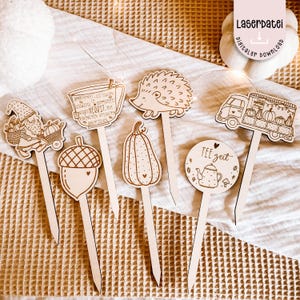 Może przedstawiać: Zestaw drewnianych dekoracyjnych patyczków z motywami jesiennymi. Patyczki przedstawiają obrazy gnoma, żołędzia, dyni, jeża, filiżanki herbaty, furgonetki z dyniami oraz koła z czajnikiem i napisem "Tee Zeit".