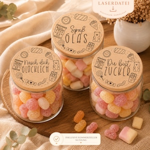 Puede incluir: Tres tarros de vidrio llenos de caramelos de goma de colores. Cada tarro tiene una tapa de madera con texto grabado e ilustraciones con temática de caramelos. El texto de las tapas dice "Nasch dich GLÜCKLICH", "Spaß OLAS" y "Du bist ZUCKER".