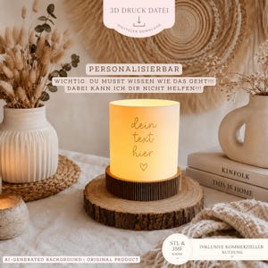 Könnte beinhalten: Eine zylindrische Lampe mit einem leuchtend weißen Schirm, der den Text "dein text hier" und ein Herzsymbol zeigt, auf einem dunkelbraunen, gerippten runden Sockel. Die Lampe steht auf einer Holzscheibe, umgeben von Vasen und Büchern.