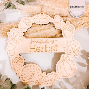 Könnte beinhalten: Ein hölzerner Herbstkranz mit lasergeschnittenen Designs von Eicheln, Blättern, Pilzen, einem Kürbis und einem Igel. Die Worte "Happy Herbst" sind auf einem Banner eingraviert. Der Kranz ist aus hellem Holz gefertigt und hat eine Aufhängeschlaufe.