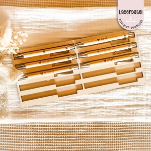 Peut inclure: Un ensemble de stylos en bois dans un support découpé au laser. Les stylos ont des détails argentés et sont gravés de mots et d'un symbole de cœur. Le support a un design géométrique. Le texte "Laserdatei digitaler download" est également visible.
