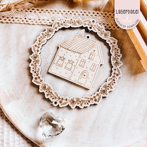 Puede incluir: Adorno de madera con forma de casa con un borde de corona floral. La casa tiene ventanas, una puerta y una chimenea. La corona está adornada con hojas y flores. El texto "Laserdatei digitaler download" es visible.