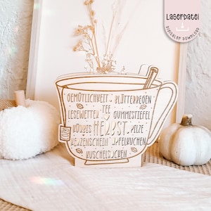 Op de afbeelding: Een houten bord in de vorm van een theekopje met Duitse woorden als "Gemütlichkeit" en "Blätterregen". Het bord heeft een gezellig thema, met een kleine pompoen en gedroogde bloemen op de achtergrond. Het bord heeft ook de tekst "Laserdatei digital download."
