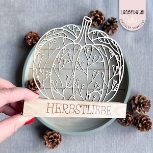 Pode incluir: Decoração de abóbora de madeira com um design detalhado e vazado. A abóbora está sobre uma base de madeira com a palavra "HERBSTLIEBE" gravada. O fundo inclui pinhas e uma bandeja azul clara.