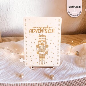 Può includere: Calendario dell'Avvento in legno con un design a schiaccianoci. Il calendario presenta le parole "magische ADVENTSZEIT" e i numeri da 1 a 24. Il design è tagliato al laser e include un piccolo supporto a forma di stella. Sfondo in tessuto bianco con ornamenti dorati.
