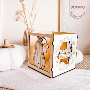 K&ouml;nnte beinhalten: Eine w&uuml;rfelf&ouml;rmige Holzlaterne mit lasergeschnittenen Herbstmotiven. Die Laterne zeigt einen K&uuml;rbis und die Worte "Happy Herbst". Das Design umfasst Bl&auml;tter, Eicheln und einen Igel. Der Text "Laserdatei digitaler download" ist ebenfalls sichtbar.