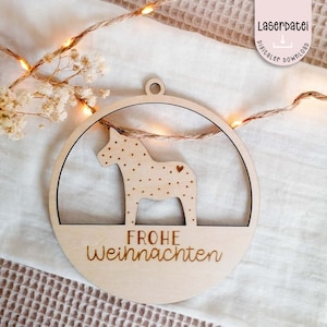 Puede incluir: Un adorno navideño de madera con un caballo Dala blanco con lunares marrones y un pequeño corazón en un lado. El adorno es redondo y tiene el texto "Frohe Weihnachten" escrito en la parte inferior.