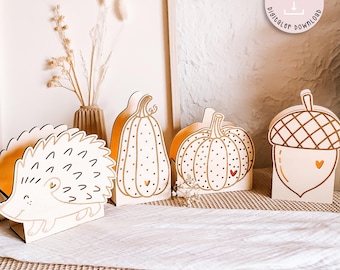 May include: A collection of wooden fall-themed decorations. Includes a hedgehog, two pumpkins, and an acorn, all with gold accents and heart details. The decorations are set on a white surface with a neutral background, suggesting a seasonal or autumn theme.