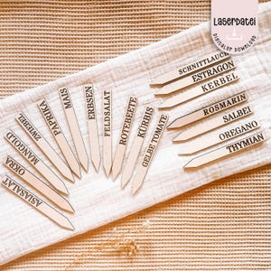 Może przedstawiać: Drewniane znaczniki ogrodowe z nazwami roślin w czarnych literach, w tym "Okra", "Asiasalat" i "Mangold". Znaczniki są ułożone na białej tkaninie. Widoczny jest tekst "Laserdatei digital download".