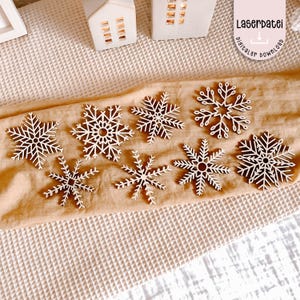 Può includere: Una collezione di sette ornamenti a forma di fiocco di neve in legno bianco con intricati disegni intagliati. I fiocchi di neve sono disposti su un tessuto beige. Due piccoli portacandele bianchi a forma di casa sullo sfondo. Il testo "Laserdatei digitaler download".