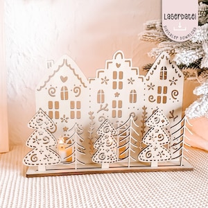 Könnte beinhalten: Eine hölzerne Weihnachtsdorf-Szene mit drei Häusern und mehreren Bäumen. Die Häuser haben ausgeschnittene Fenster und dekorative Details. Die Bäume sind ebenfalls mit aufwendigen Mustern ausgeschnitten. Eine Kerze brennt hinter den Bäumen. Der Text "Laserdatei digitaler download" ist sichtbar.