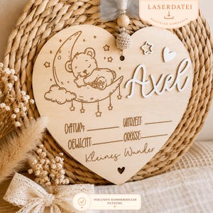 May include: Wooden heart-shaped sign with the name "Axel" in white script. The sign features a laser-etched design of a sleeping bear on a crescent moon, stars, and text fields for baby's details. The sign is a digital download.