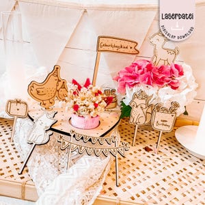 Könnte beinhalten: Ein hölzernes Tortenaufsatz-Set zum Geburtstag mit verschiedenen Tier- und Banner-Ausschnitten. Das Set enthält ein Reh, einen Fuchs, ein Huhn und eine Katze sowie ein Banner mit der Aufschrift "Happy Birthday". Ein kleiner rosa Behälter enthält Trockenblumen.